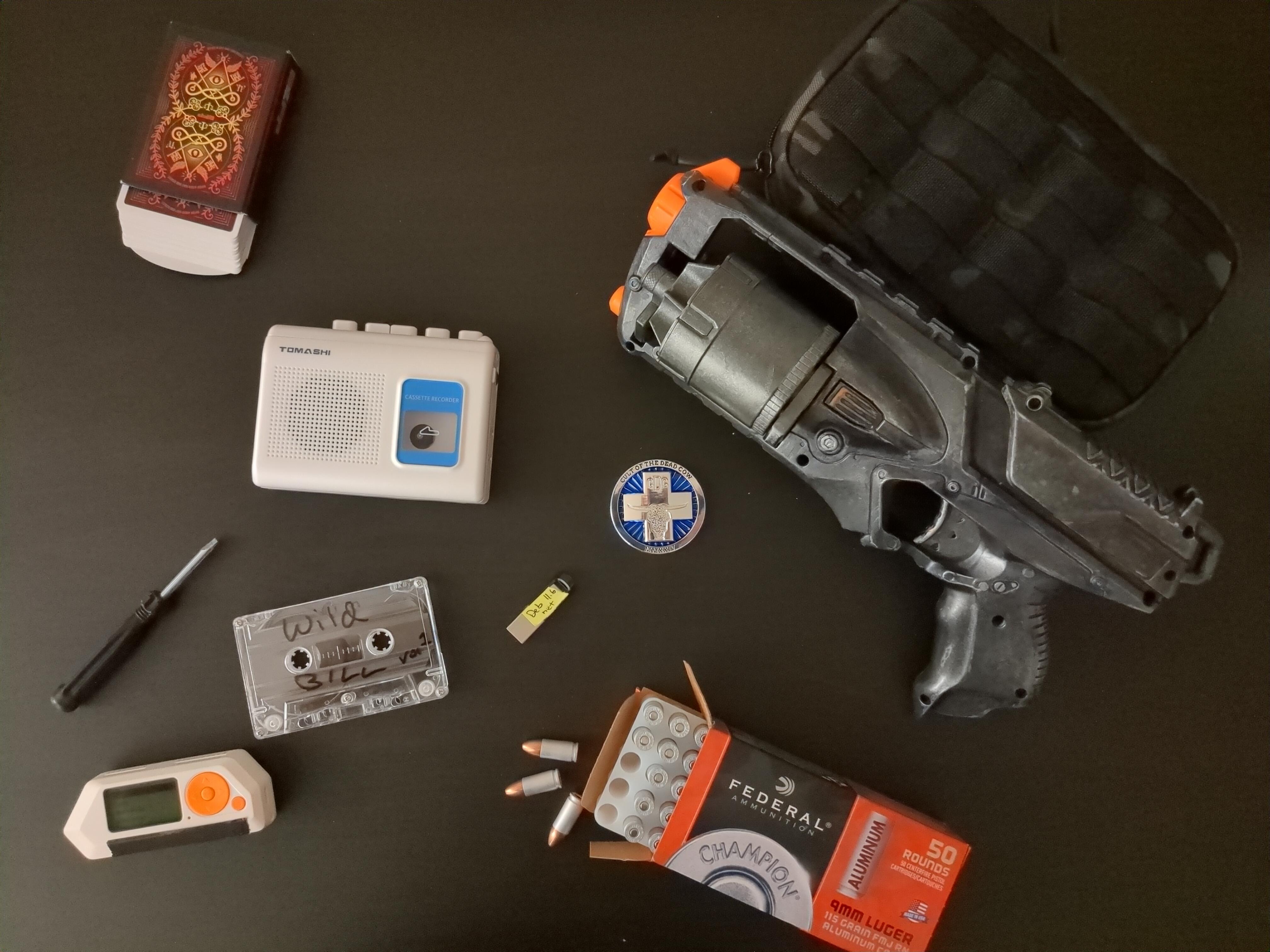 an overhead tableau of items scattered on a desktop: a cassette player, a cassette labeled Wild Bill Vol 1, a box of pistol ammunition and several loose cartridges, a silver NERF blaster, a deck of playing cards, a challenge coin, a USB thumb drive